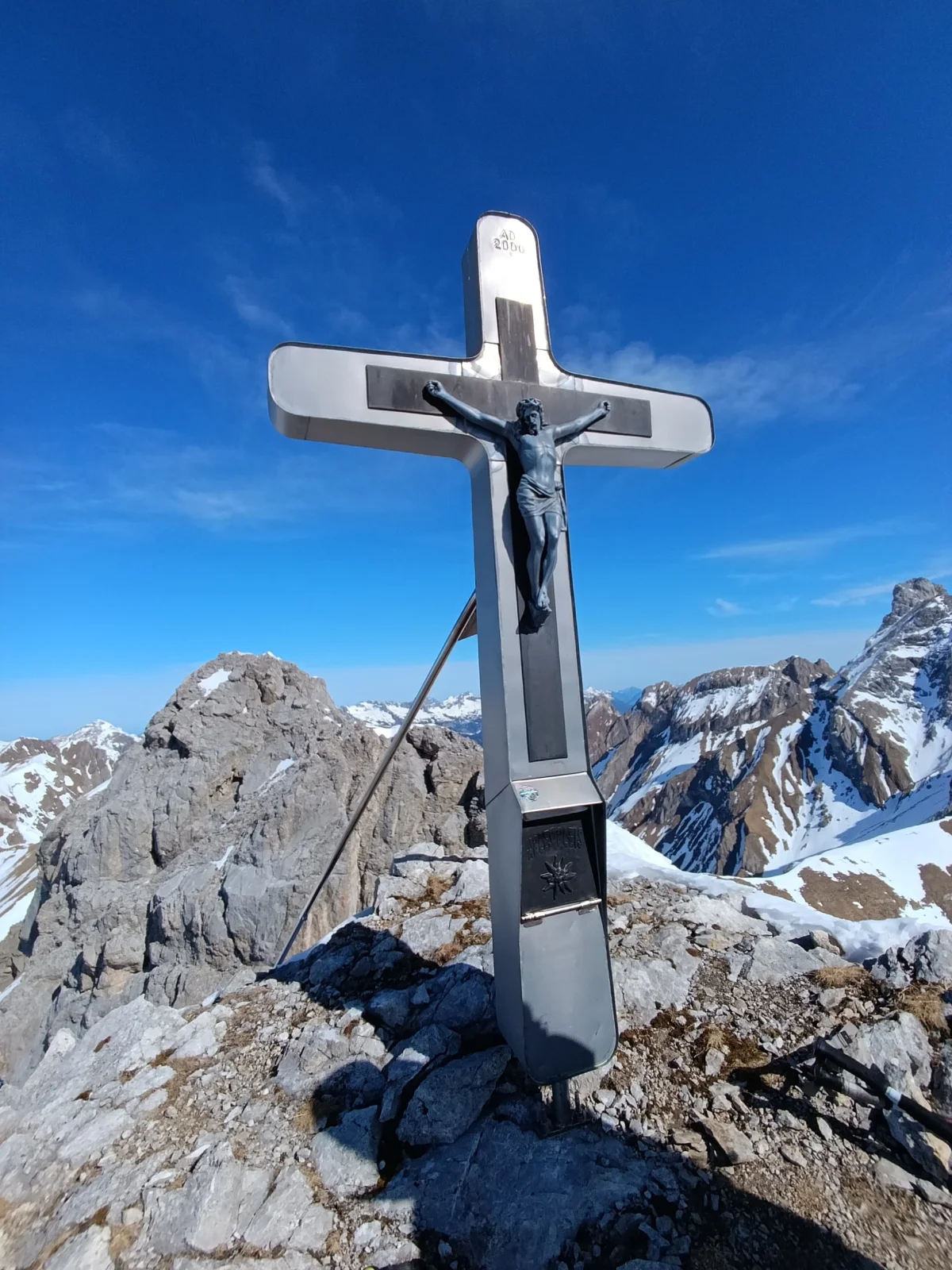 Gipfelkreuz der Aples-Pleisspitze vor verschneiten Bergen und blauem Himmel.