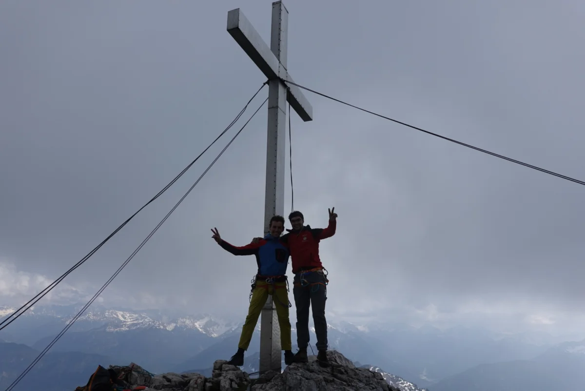 Zwei Bergsteiger am Gipfelkreuz der Gehrenspitze unter wolkigem Himmel.
