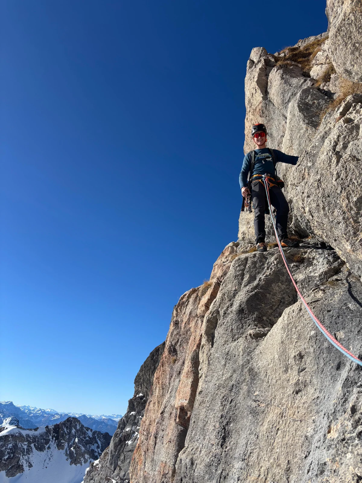Kletterer mit Helm und Kletterausrüstung auf einem Felsabsatz vor blauem Himmel.