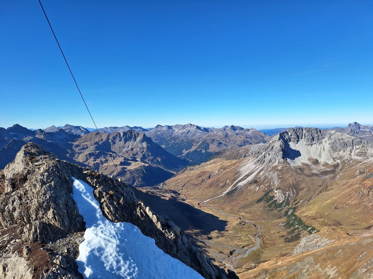 Narrow rocky ridge with snow and panoramic views of the surrounding mountains.