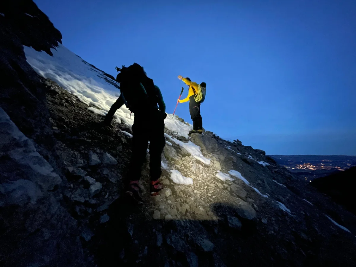 Zwei Bergsteiger mit Stirnlampen im Morgengrauen auf einem verschneiten Grat, unten die Lichter des Tals.