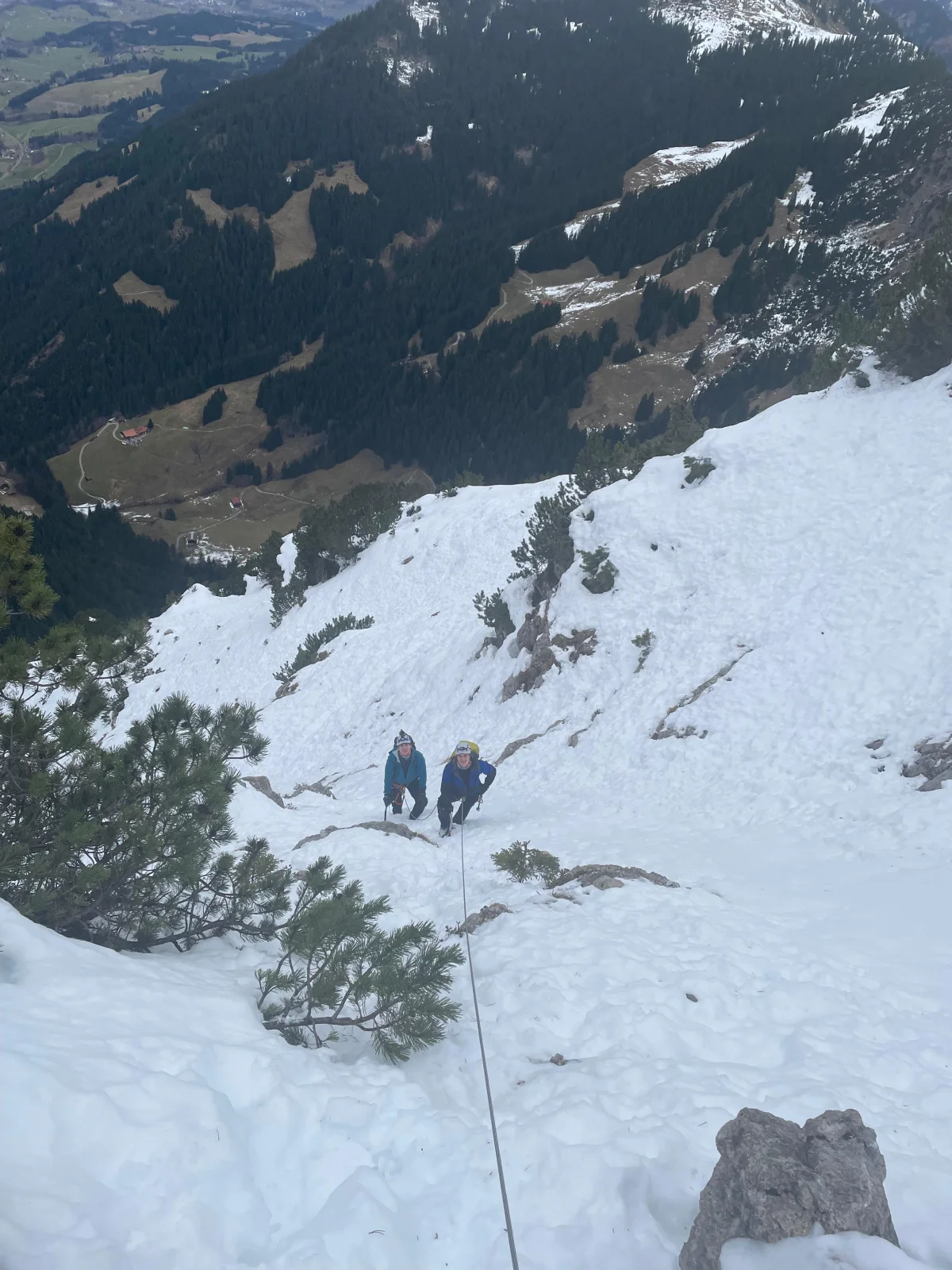 Zwei angeseilte Bergsteiger im Schneefeld unterhalb der Nordwand, im Hintergrund Wald und Tal.